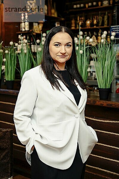 A confident woman with dark hair wearing a white blazer and black dress stands in front of a bar