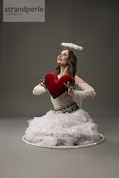 Young female sitting in studio on gray background wearing white dress hold red heart