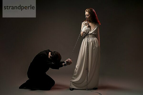 Full body of young woman in white dress holding chain tied around hands of kneeling man in black suit