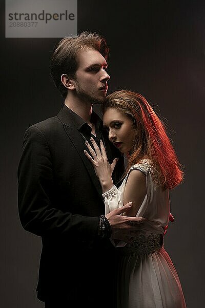 Confident young man in black suit embrace woman in bridal dress while standing on black background