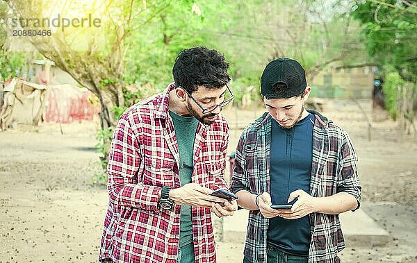 Two teenage guys pointing and checking their cell phones  Two friends looking at the content of their cell phone. Friend showing cell phone to his friend  Smiling friends checking cell phones