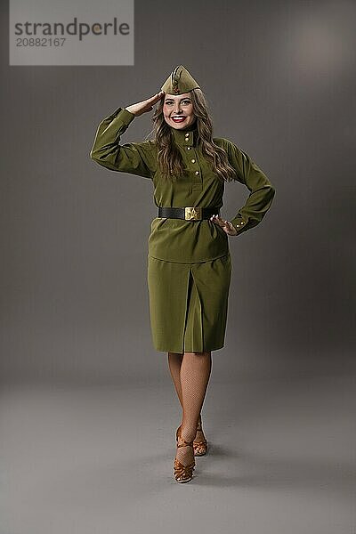 Full length of confident young military female in green uniform of USSR army and hat with long hair smiling and looking at camera while saluting