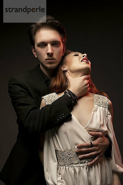 Confident young man in black suit strangling woman in bridal dress while standing on black background