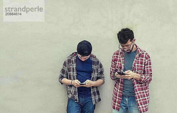 Two teenage friends on a wall checking their cell phones  Two friends leaning on a wall texting on their phones. Friend showing cell phone to his friend  Smiling friends checking cell phones