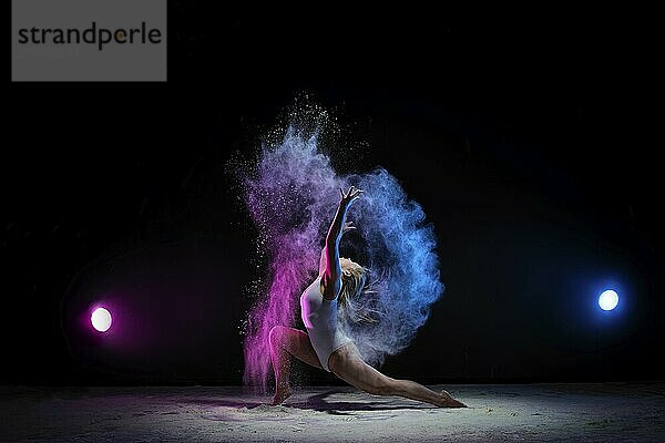 Female sexy gymnast in beige bodysuit bending gracefully with her hands up on black background with colored dust all over body shot made with color filters