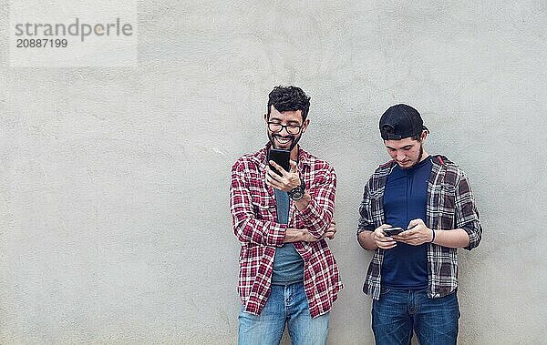 Two smiling friends leaning on a wall checking their cell phones  Friends leaning on a wall texting on their phones. Friend showing cell phone to his friend  Smiling friends checking cell phones