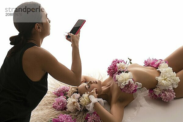 High angle of woman with dark hair using smartphone with black screen to take photo of female model with fresh flowers on breast and hips