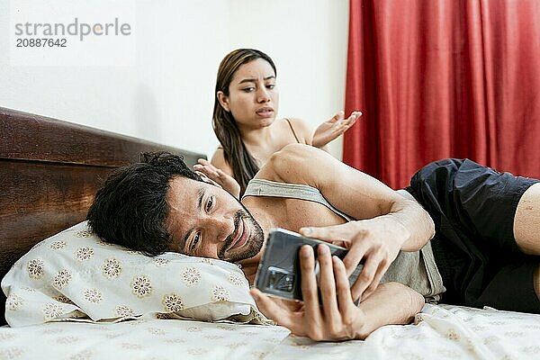 Husband with cell phone ignoring wife in bed. Annoyed woman with husband while holding cell phone in bed. Cheating Husband Texting On Phone Ignoring Wife Lying In Bed