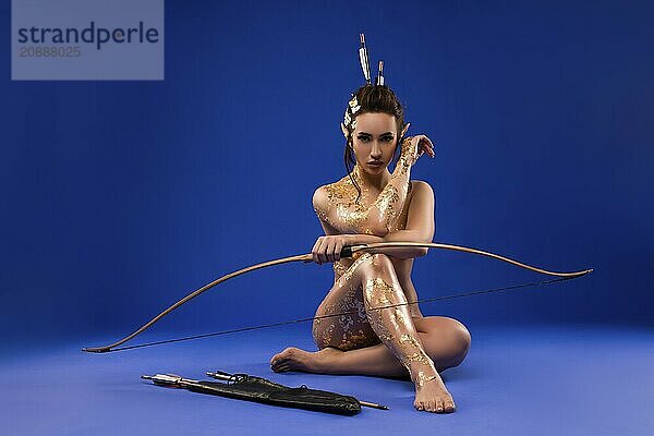 Full length of sensual confident female model with golden paint on naked body and elf ears sitting on floor arrows and bow  blue background