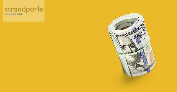 Roll of one hundred dollar bills secured with a rubber band on a yellow ochre background