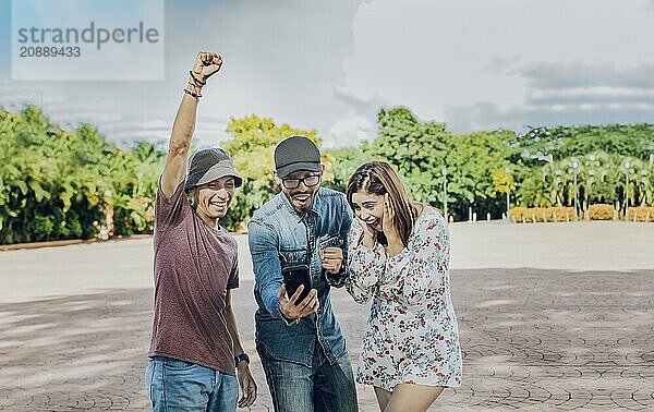 Three cheerful friends looking at cell phone making gesture winner outdoors. Excited teenage friends looking at cell phone screen on the street