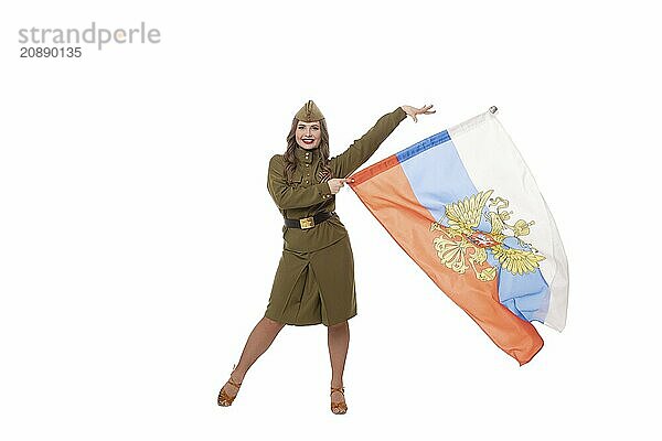 Positive female model with long wavy hair in military uniform and hat smiling brightly and looking at camera while demonstrating Russian flag against white background