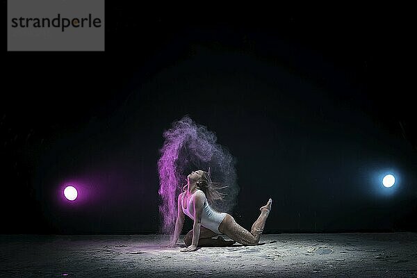 Female sexy gymnast in beige bodysuit bending gracefully on black background with colored dust all over body shot made with color filters