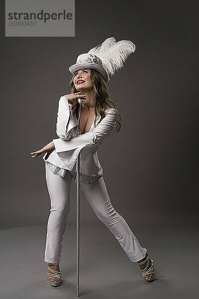 Full body of smiling young female in art deco suit with feathers on hat leaning on white cane standing against gray background looking up