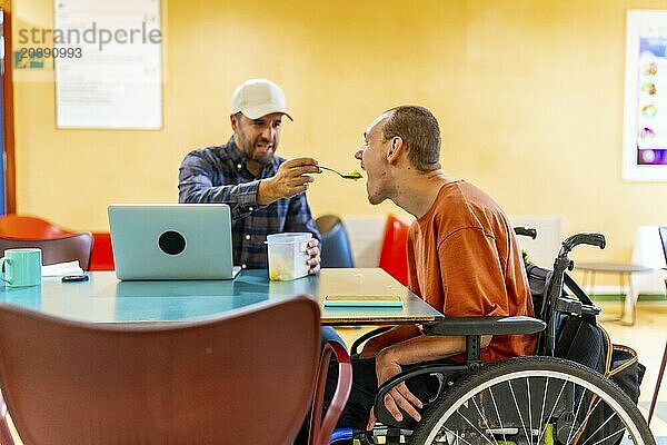 Colleague feeding a man with cerebral palsy in a coworking cafeteria in a modern space