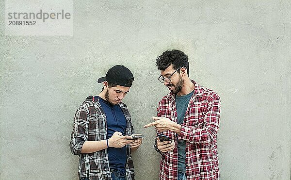 Two young friends leaning against a wall checking their cell phones  Two friends leaning against a wall looking at the content on their phones. Friend showing cell phone to his friend outdoors