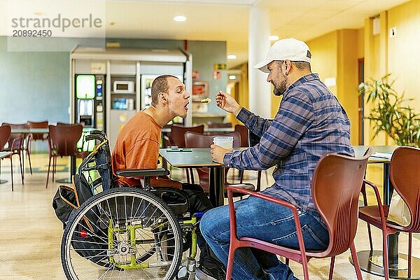 Adult caucasian and casual man feeding a colleague with disability in a coworking cafeteria