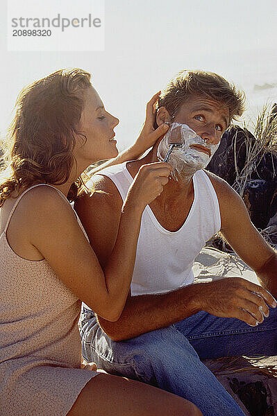 Close up of young couple at the beach. Woman wet shaving a man.