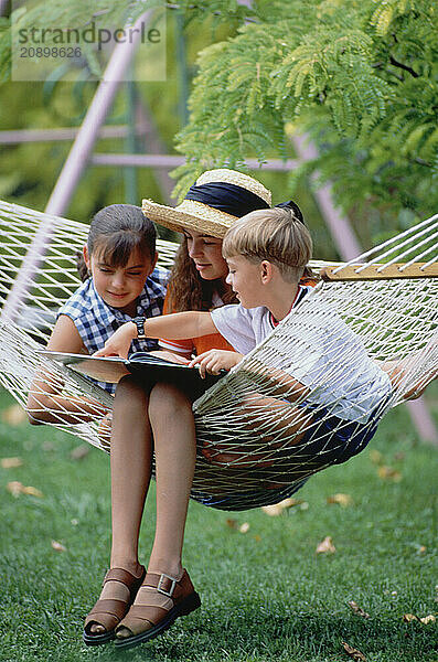 Three children. Two girls & boy outdoors in hammock reading.