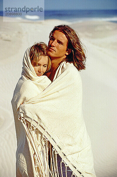 Young couple wrapped in blanket  standing on a beach.