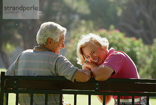 Back view close up of senior man & woman couple sitting outdoors on park bench.