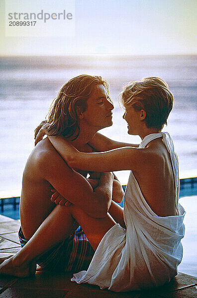 Young couple sitting closely together by oceanside pool at sunset.