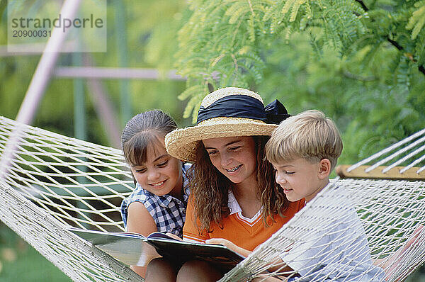 Three children. Two girls & boy outdoors in hammock reading.