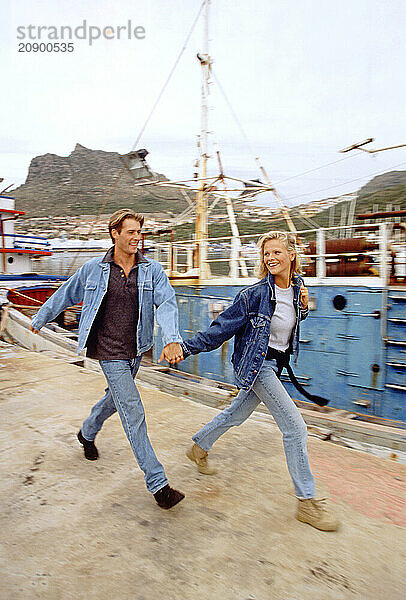 South Africa. Cape Town. Motion impression of young couple walking on quay by boats in harbour.