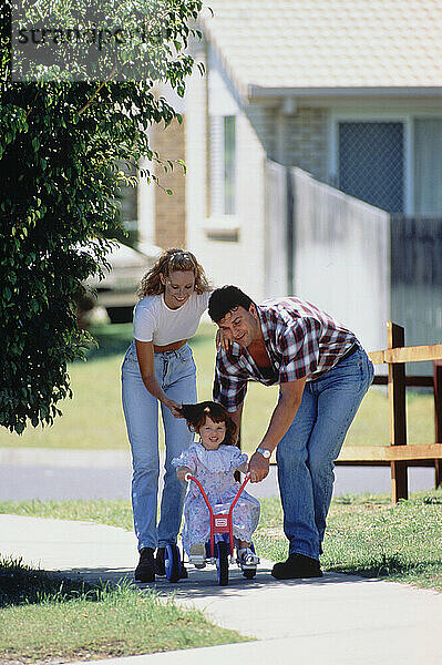 Family. Parents helping little daughter to ride tricycle outside on pavement.