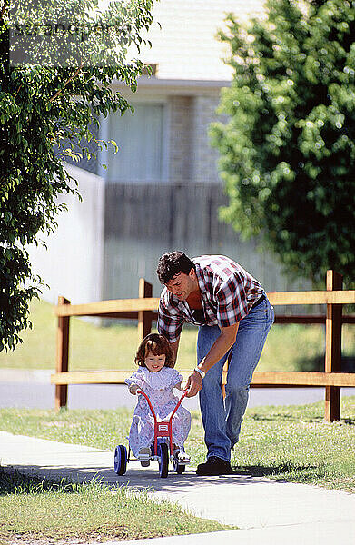 Family. Father helping little daughter to ride tricycle outside on pavement.