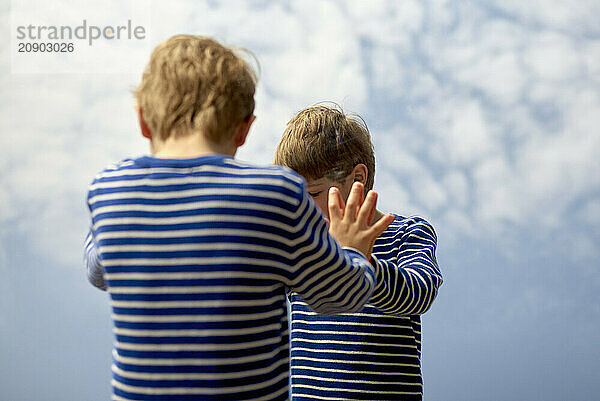 Two young children in striped shirts stand against a backdrop of clouds  with one comforting the other by placing a hand on their shoulder.