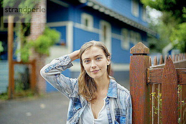 Smiling young woman standing by a wooden fence with a blue house in the background.