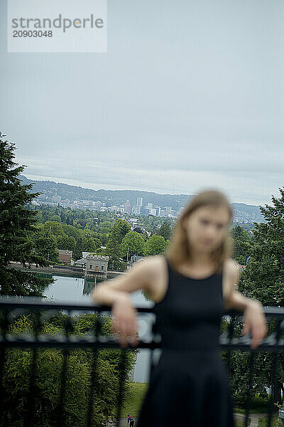 A female in black athletic wear rests on a railing with a blurred city park landscape in the background.