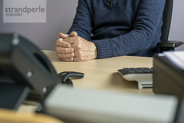 Close-up of a person's clasped hands resting on a desk in an office setting with a blurred foreground.