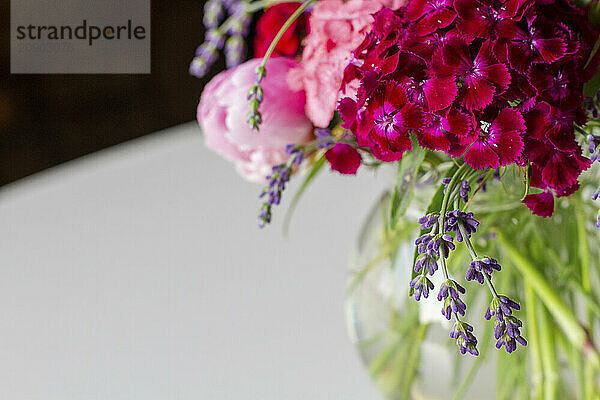 Vibrant bouquet of pink peonies and purple flowers in a clear vase on a white surface with a blurred background.