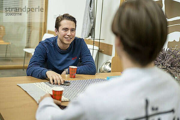 Smiling young man sitting at a table indoors having a conversation with an out-of-focus person sitting across from him.