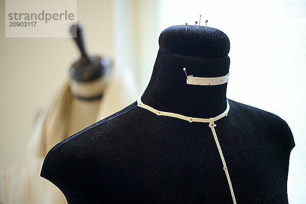 Black dress form with measuring tape on the neck  pins on top  and a blurred background featuring a kitchen knife block.