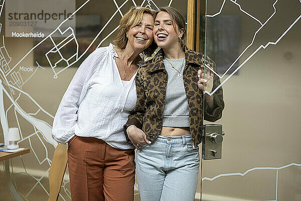 Smiling woman with her arm around a younger woman  both standing in an office with a glass door and map design in the background.