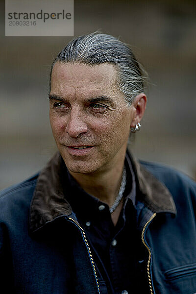 Mature man with grey hair and a leather jacket looks contemplatively to the side against a blurred background.