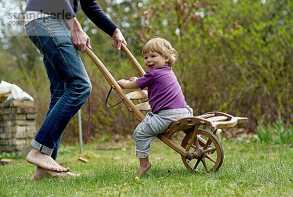 Toddler enjoys a ride in a wooden wagon outdoors  pulled by an adult on a sunny day.