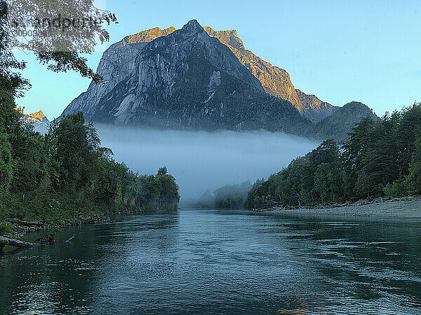 Sunrise illuminates a majestic mountain peak above a tranquil river veiled in morning mist  Golfo de Peno s Patagonia  Chile