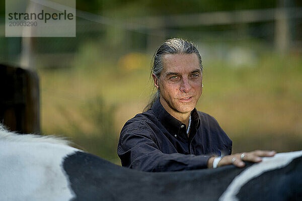 Smiling woman with long gray hair resting her arm on a horse's back in nature.