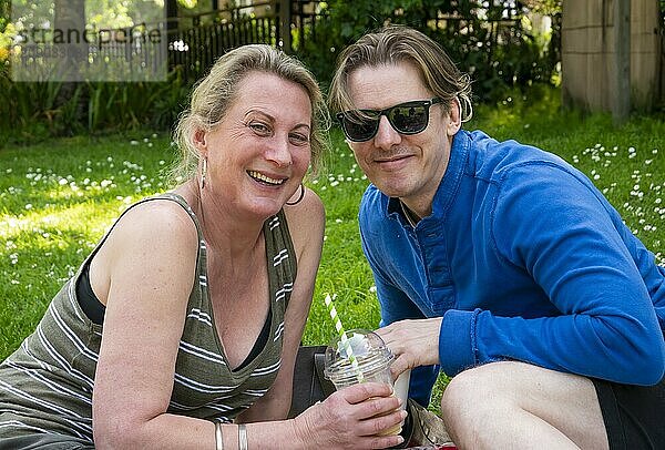 Smiling woman and man sitting on the grass enjoying a sunny day outdoors  with the man holding a drink.