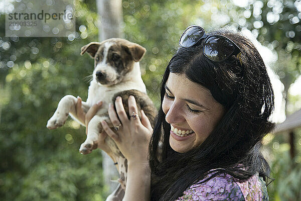 Smiling woman holding a playful puppy outdoors with trees in the background.