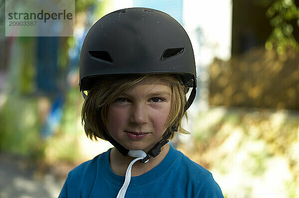 A young child wearing a black helmet and blue shirt smiles subtly outdoors with greenery and a colorful background softly blurred.