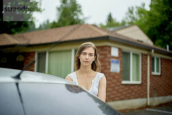 Young woman standing in front of a brick house with trees  partially obscured by a car in the foreground.