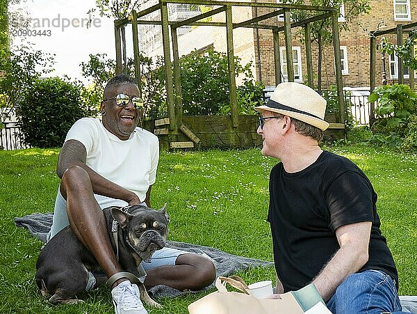 Two men and a dog enjoy a picnic on a sunny day in a grassy backyard.