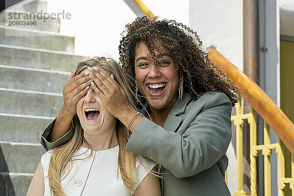 Woman covers friend's eyes with her hands as they both laugh on outdoor steps.