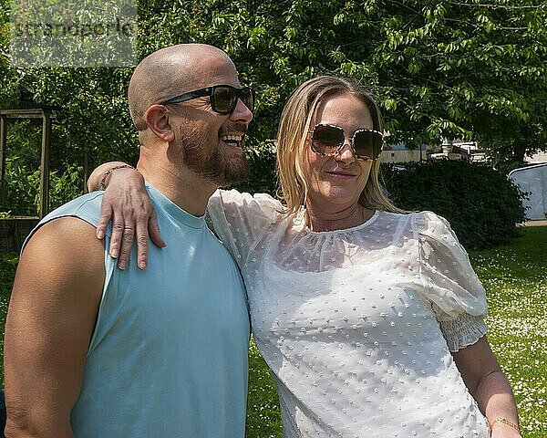 Smiling man in a blue top and sunglasses embraces a woman in a white dress  standing outdoors on a sunny day.