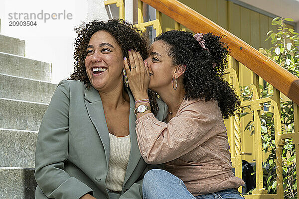 Two women sitting on steps sharing a secret  one is whispering into the other's ear and both are smiling.